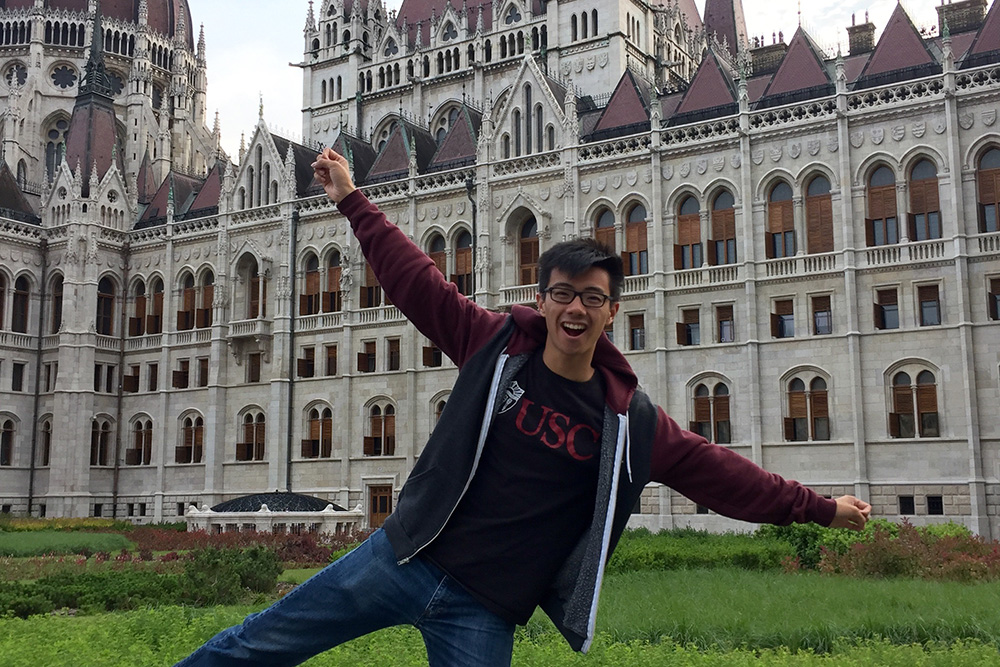 USC student Russell Yue in a USC T-shirt posing energetically in front of the Hungarian Parliament building in Budapest, Hungary, May 19, 2016.