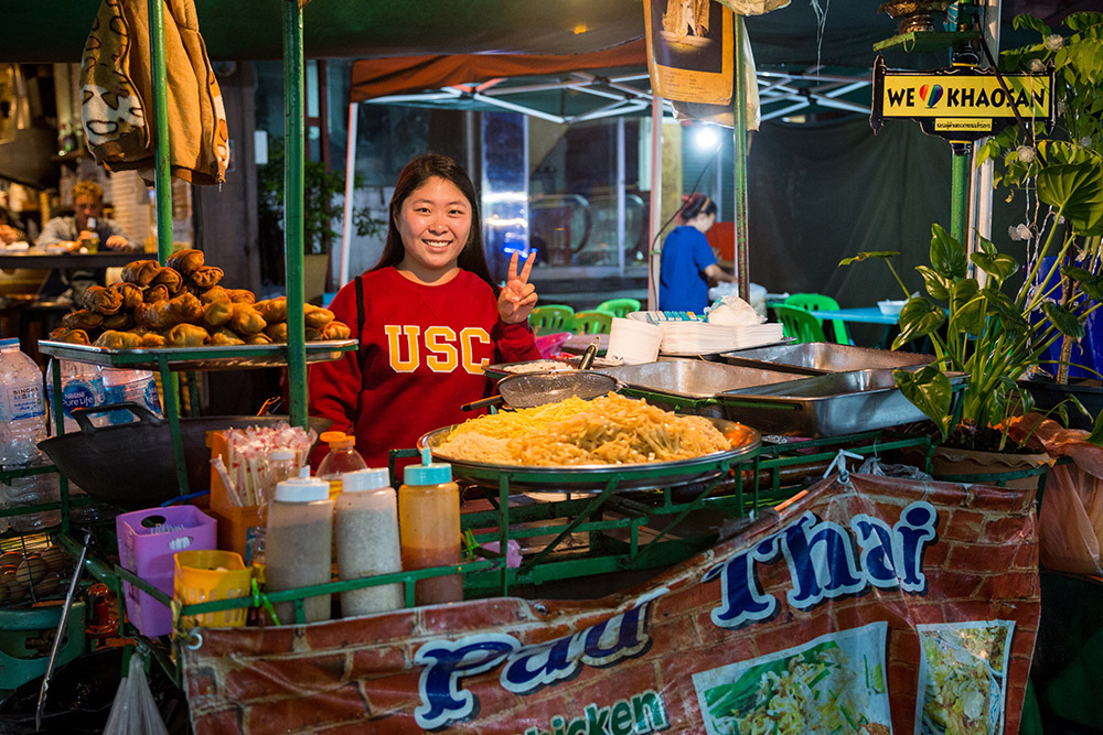 USC student at Khao San Road street food stall in Bangkok, representing university’s Student Travel Abroad programs and Student Health Insurance requirements.